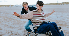 Twee vrolijke vrienden op het strand. De één duwt de rolstoel van de ander.