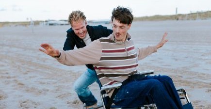 Twee vrolijke vrienden op het strand. De één duwt de rolstoel van de ander.