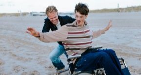 Twee vrolijke vrienden op het strand. De één duwt de rolstoel van de ander.