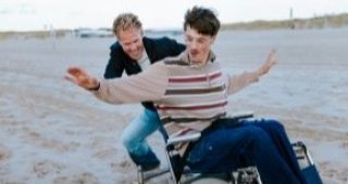 Twee vrolijke vrienden op het strand. De één duwt de rolstoel van de ander.